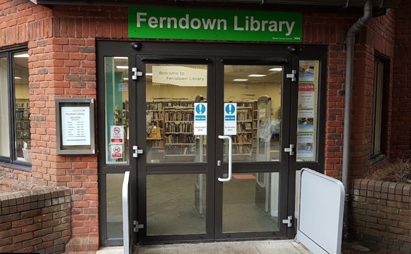 The front entrance of Ferndown Library: automatic glass doors under a green sign reading &ldquo;Ferndown Library&rdquo;, set in a red brick frontage, with bookshelves visible inside.